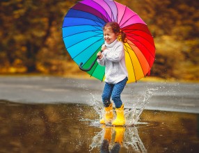 Kleines Mädchen spielt mit gelben Gummistiefeln und einem Regenschirm in den Farben des Regenbogens in einer Pfütze